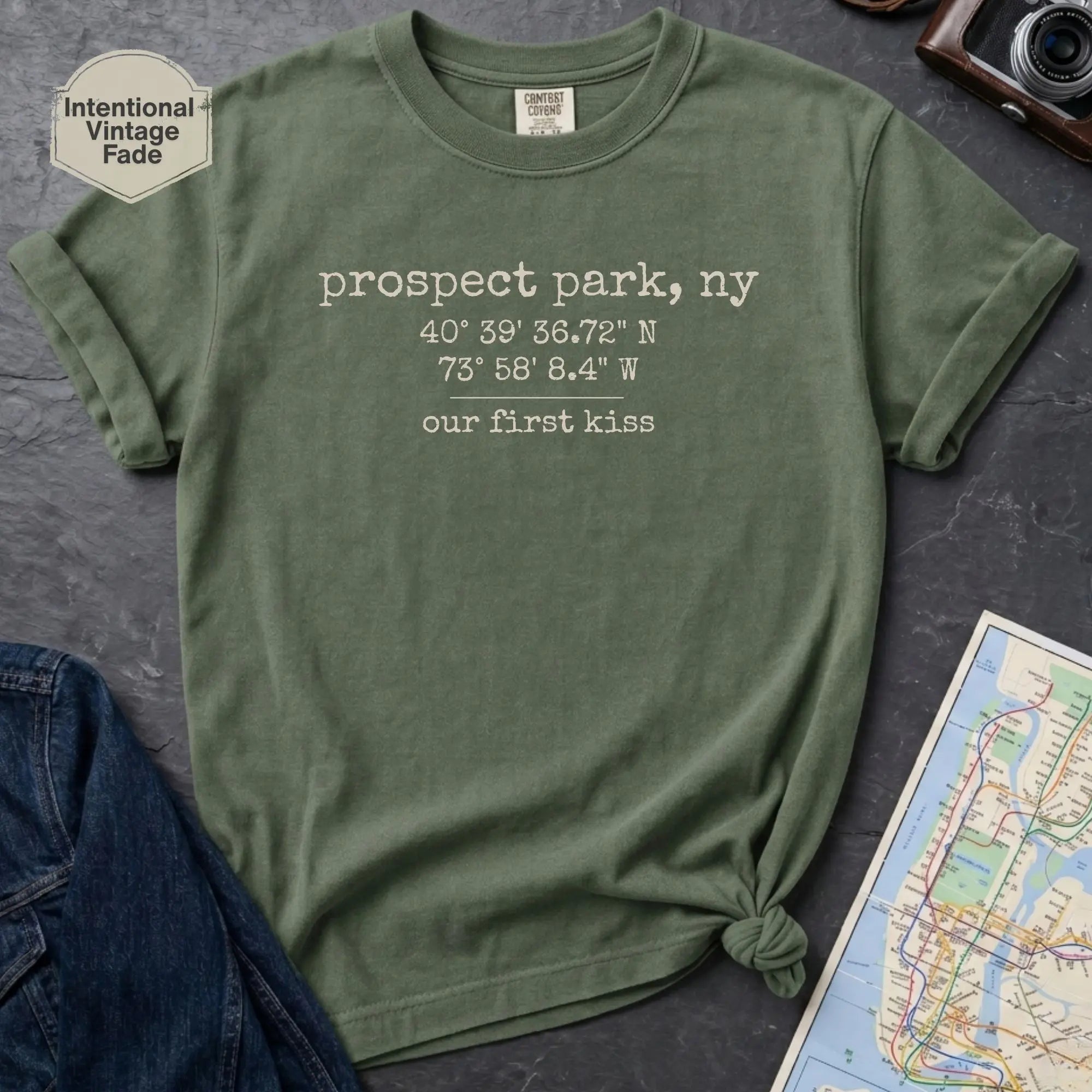 Moss colored t-shirt with text 'prospect park our first kiss' on a dark surface with a map and denim jacket.