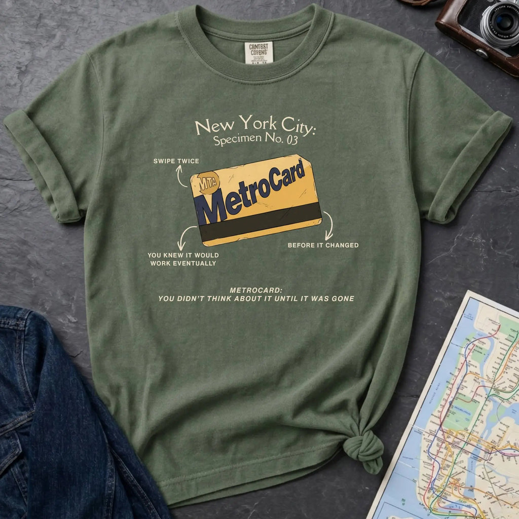 Moss green t-shirt with New York City MetroCard graphic on a gray surface with a map and jacket.