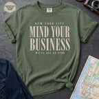 Green t-shirt with 'New York City Mind Your Business We'll All Be Fine' text on a dark surface with a map and jeans.