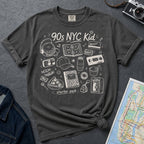 Pepper-colored Comfort Colors t-shirt with '90s NYC Kid' graphic design on a textured surface with a map and camera.