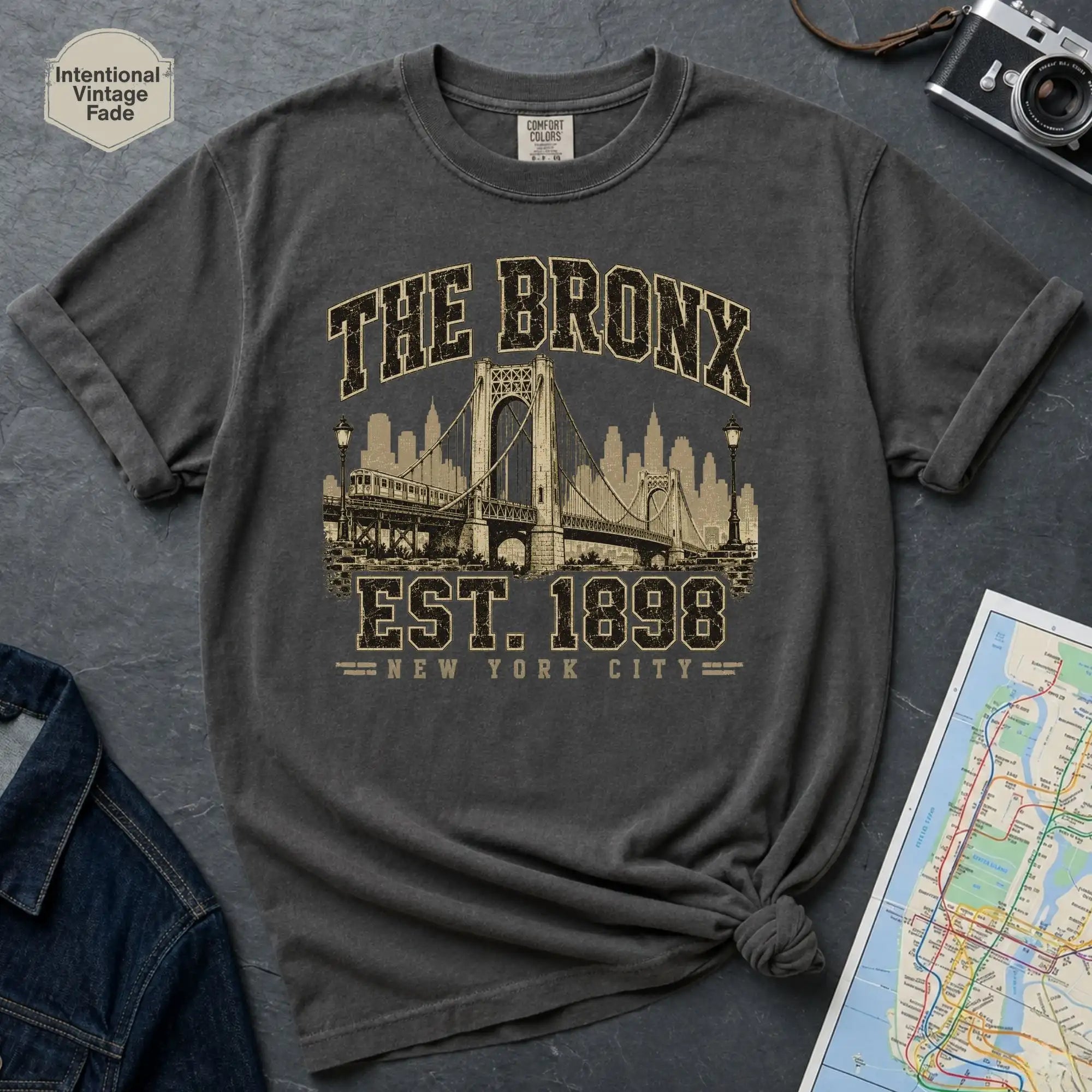 Pepper colored t-shirt with 'The Bronx Est. 1898' design on a textured surface with a map and camera.
