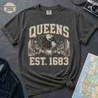 Pepper colored t-shirt with 'Queens Est. 1683' graphic design on a dark surface with a denim jacket and a map.