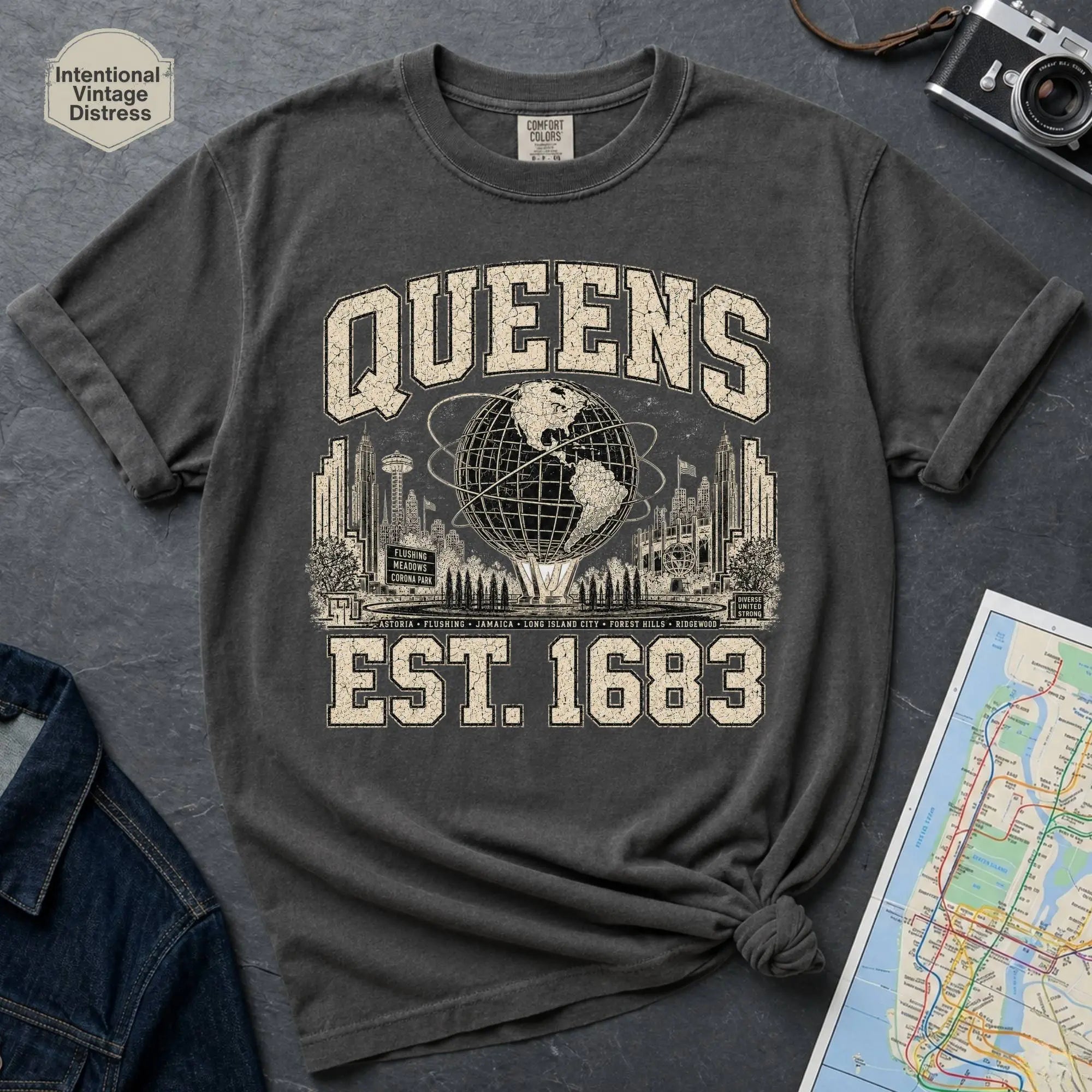 Pepper colored t-shirt with 'Queens Est. 1683' graphic design on a dark surface with a denim jacket and a map.