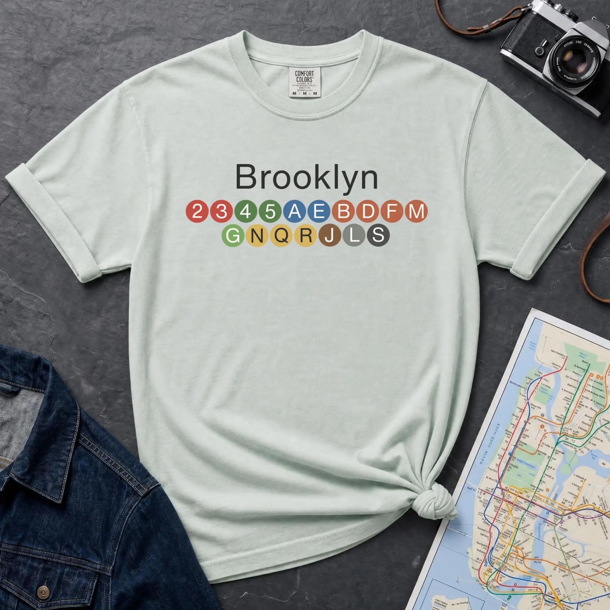 Bay colored t-shirt with 'Brooklyn' and subway line numbers on a dark surface with a map and camera.