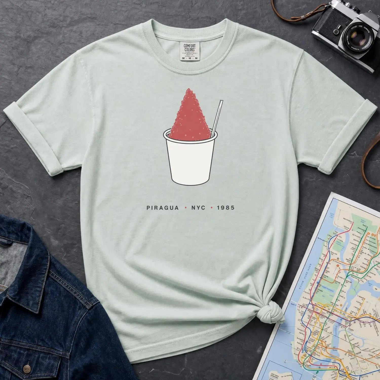 Bay colored Comfort Colors t-shirt with a piragua graphic and text, placed on a dark surface with a map and denim jacket.