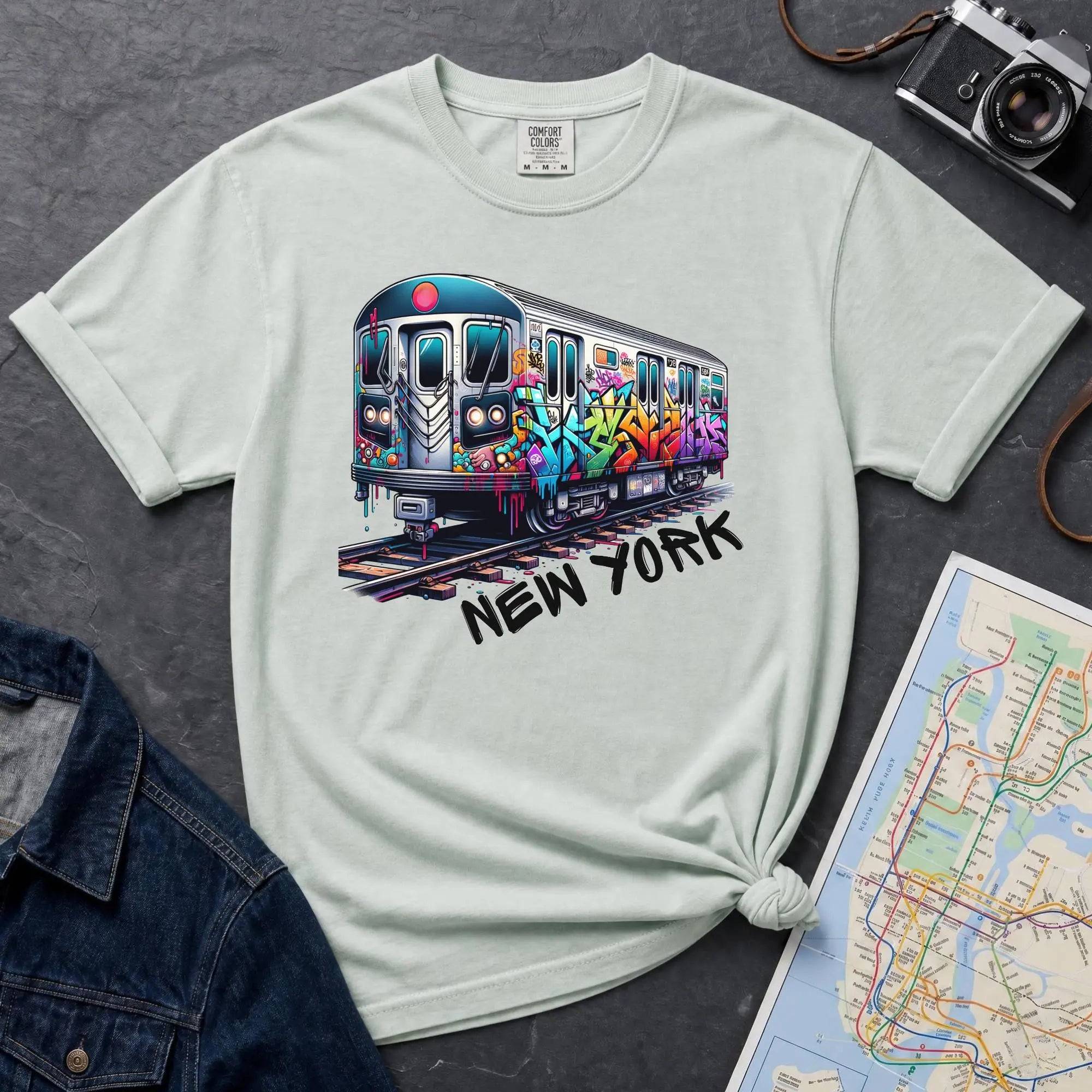 Bay colored t-shirt with a colorful train graphic and 'New York' text on a gray surface with a map and denim jacket.