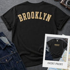 Black Comfort Colors t-shirt with 'Brooklyn' printed on the back and 'Brooklyn, New York City', on a dark surface. 
