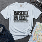 Light blue t-shirt with 'Raised in New York City' text on a dark surface with a jean jacket and a map.
