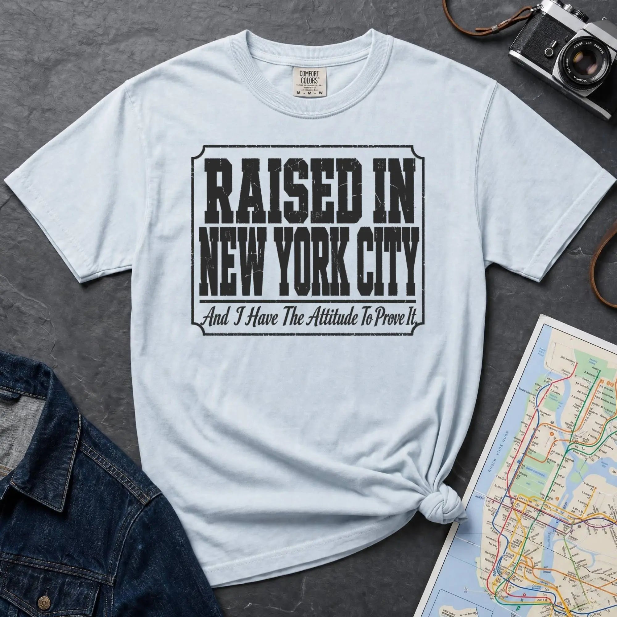Light blue t-shirt with 'Raised in New York City' text on a dark surface with a jean jacket and a map.