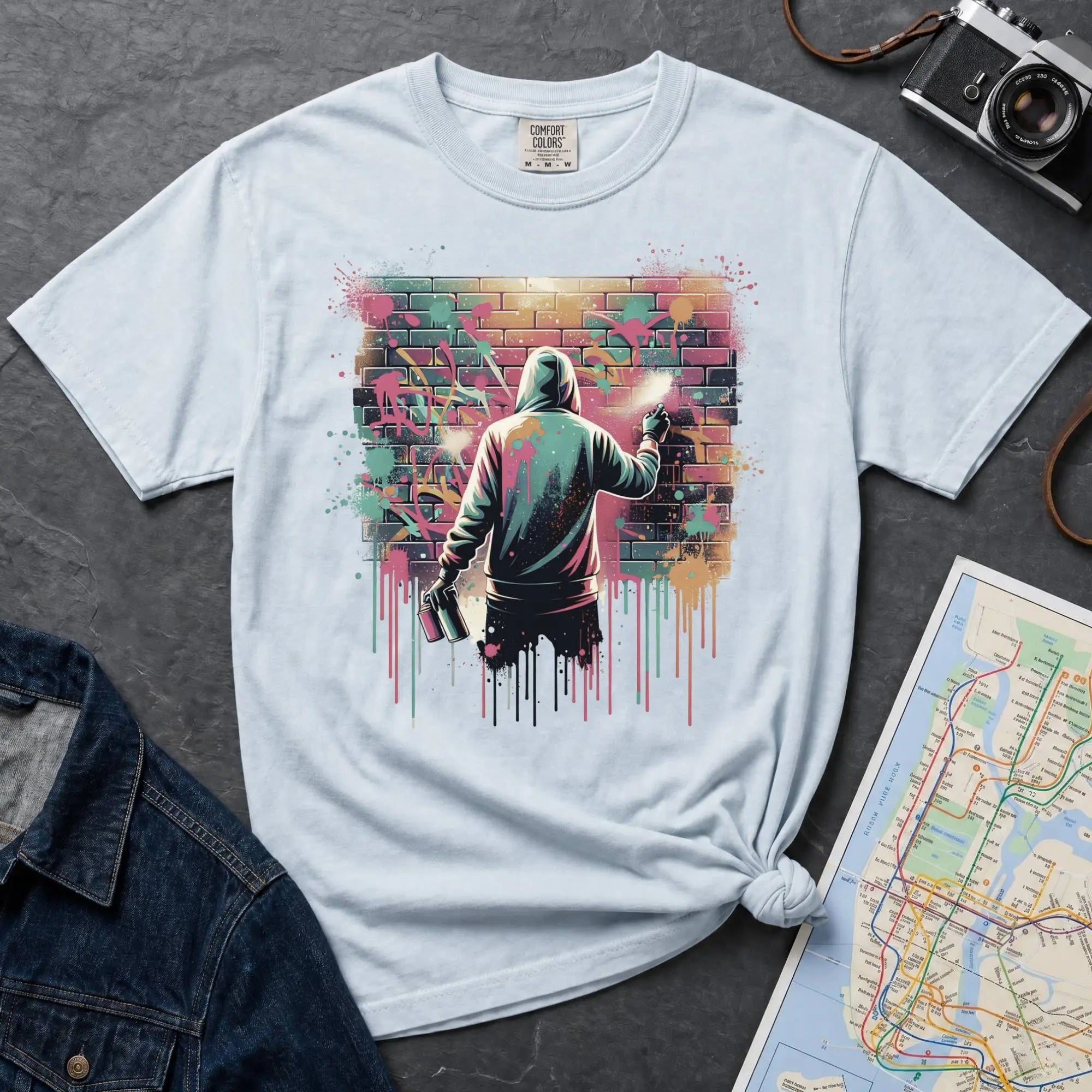 Light blue colored t-shirt with a graphic of a person spray painting a mural on a brick wall, placed on a dark surface with a denim jacket and map in the background.