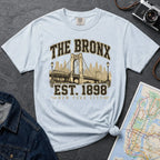 Light blue t-shirt with 'The Bronx Est. 1898' design on a gray surface with a map and camera.