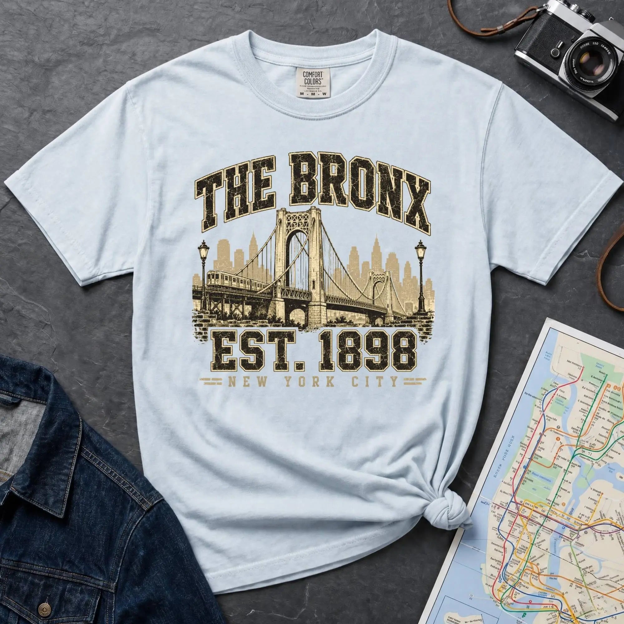 Light blue t-shirt with 'The Bronx Est. 1898' design on a gray surface with a map and camera.
