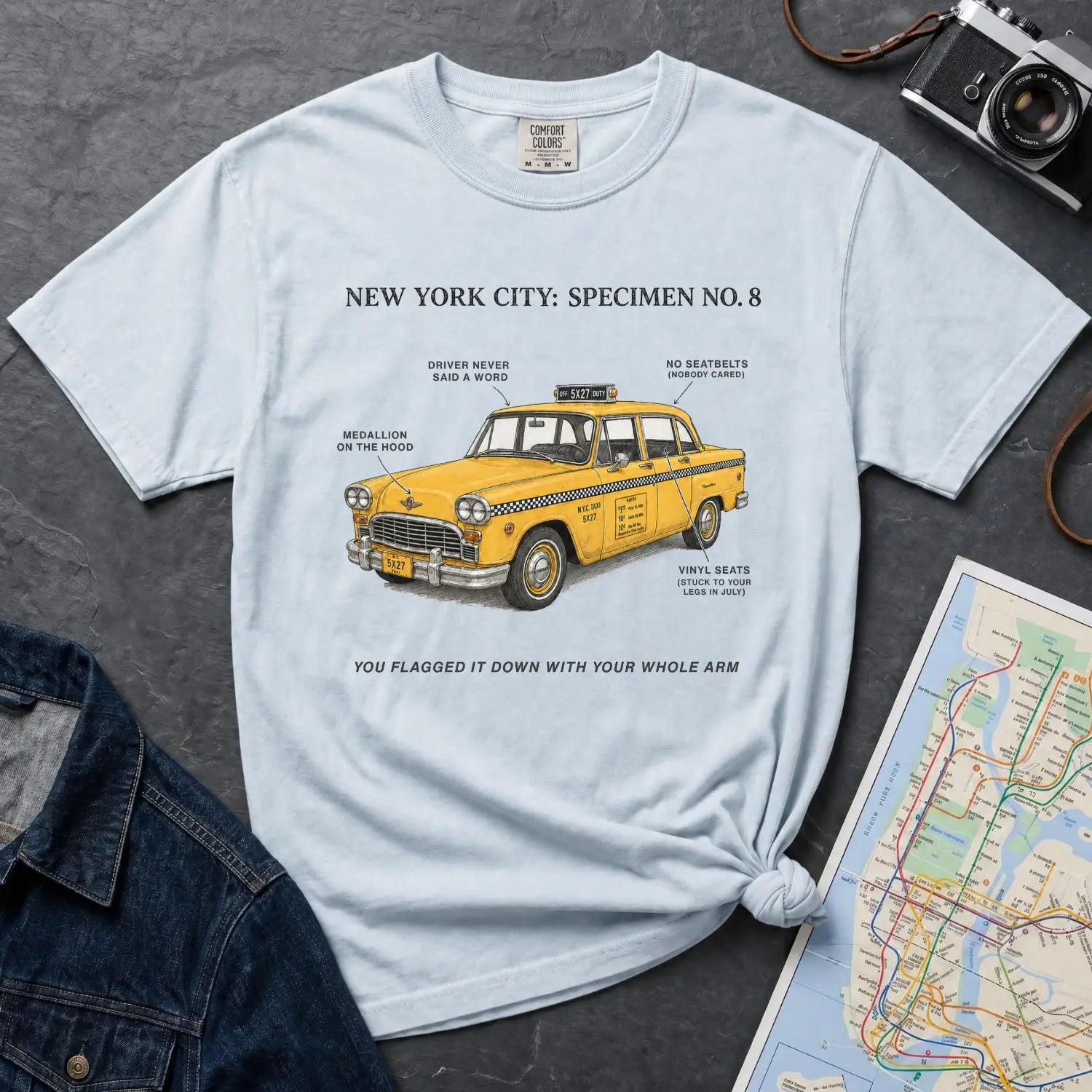 Light blue Comfort Colors t-shirt with yellow taxi graphic and text on a dark surface with denim jacket and map.