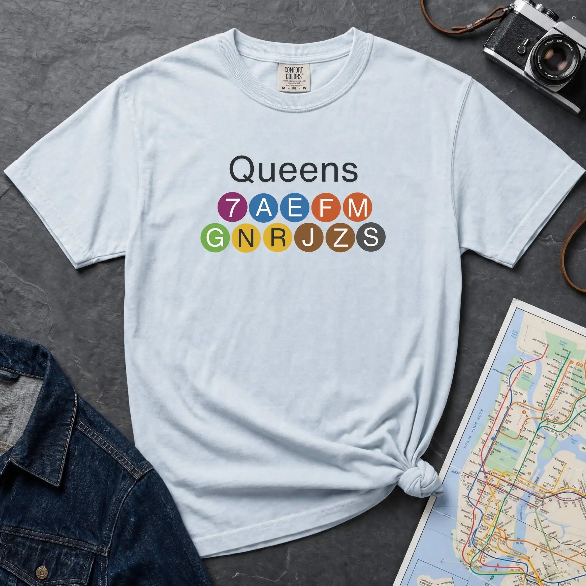 Light blue t-shirt with 'Queens' and NYC subway lines on a dark surface with a map and denim jacket.