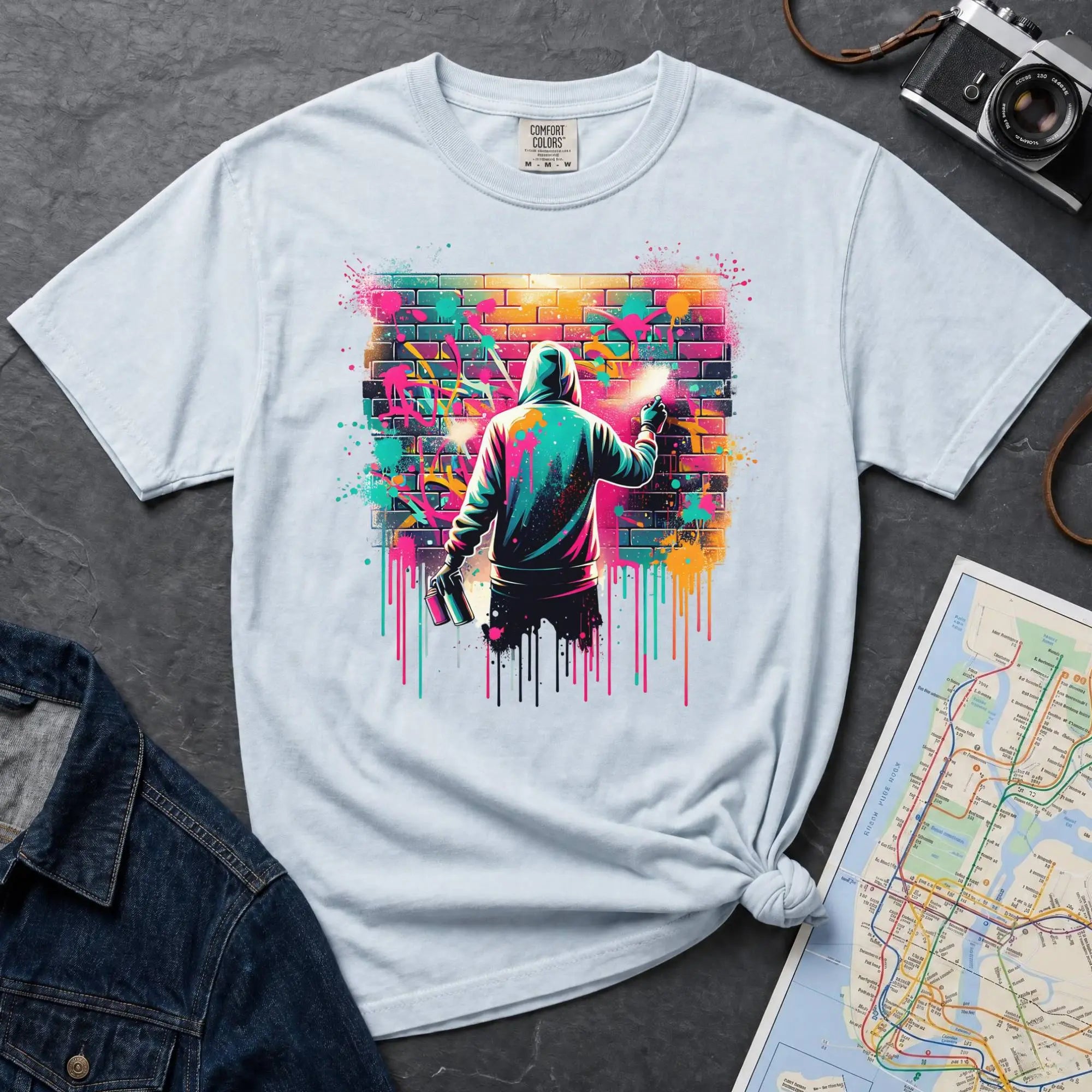 Light blue t-shirt with a graphic of a person spray painting a colorful mural on a brick wall.