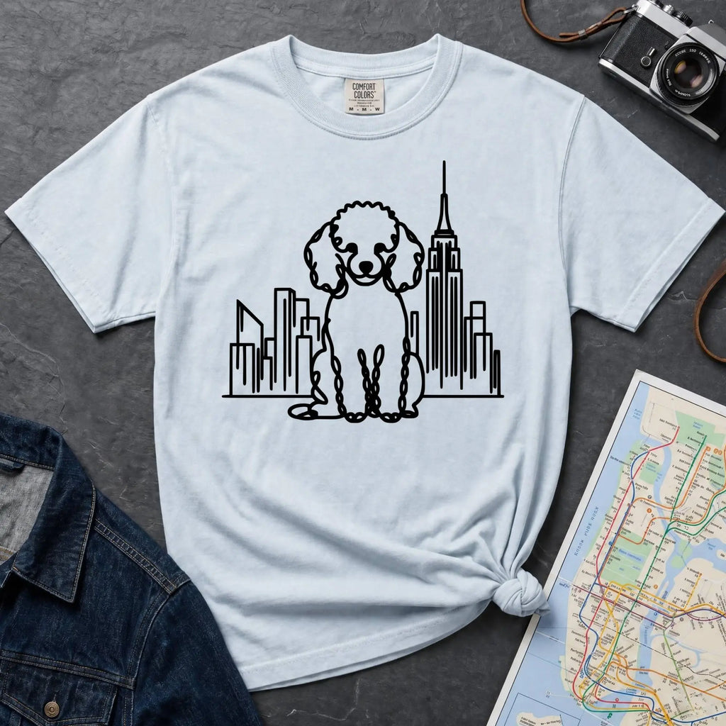 Light blue t-shirt with a poodle and cityscape design on a dark surface with a map and camera.