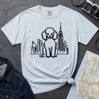 Light blue t-shirt with a poodle and cityscape design on a dark surface with a map and camera.