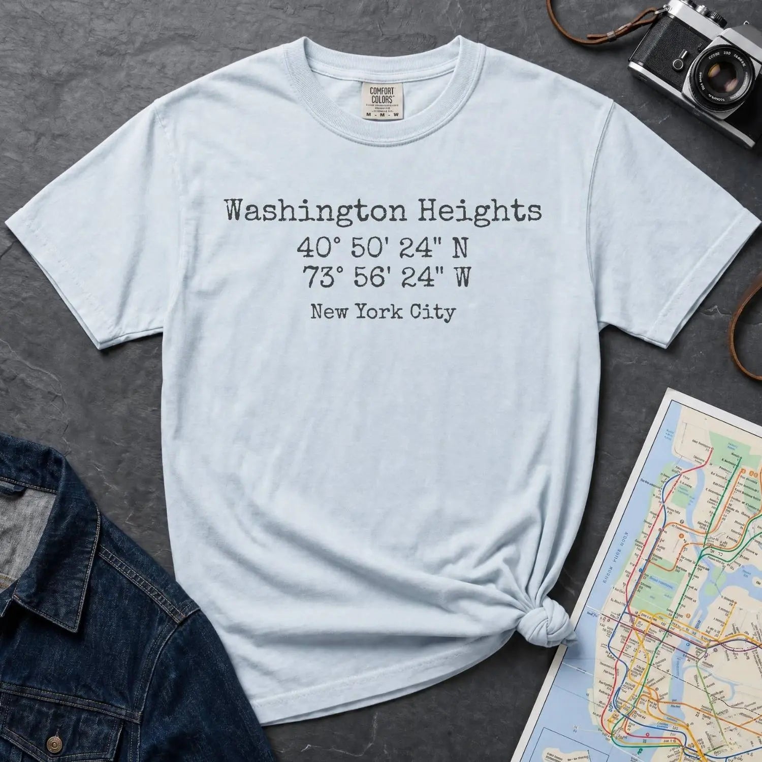 Light blue colored Comfort Colors t-shirt with 'Washington Heights' text with coordinates and 'New York City' on a dark surface with a map and denim jacket.