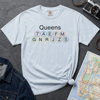 Light blue t-shirt with 'Queens' and subway line numbers on a dark surface with a map and camera.