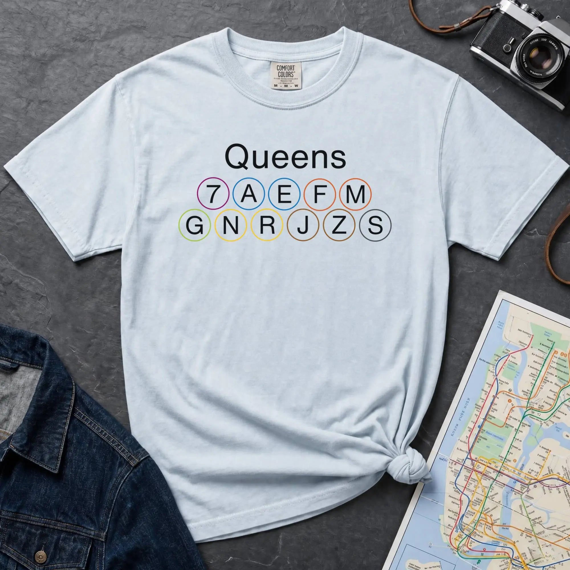 Light blue t-shirt with 'Queens' and subway line numbers on a dark surface with a map and camera.