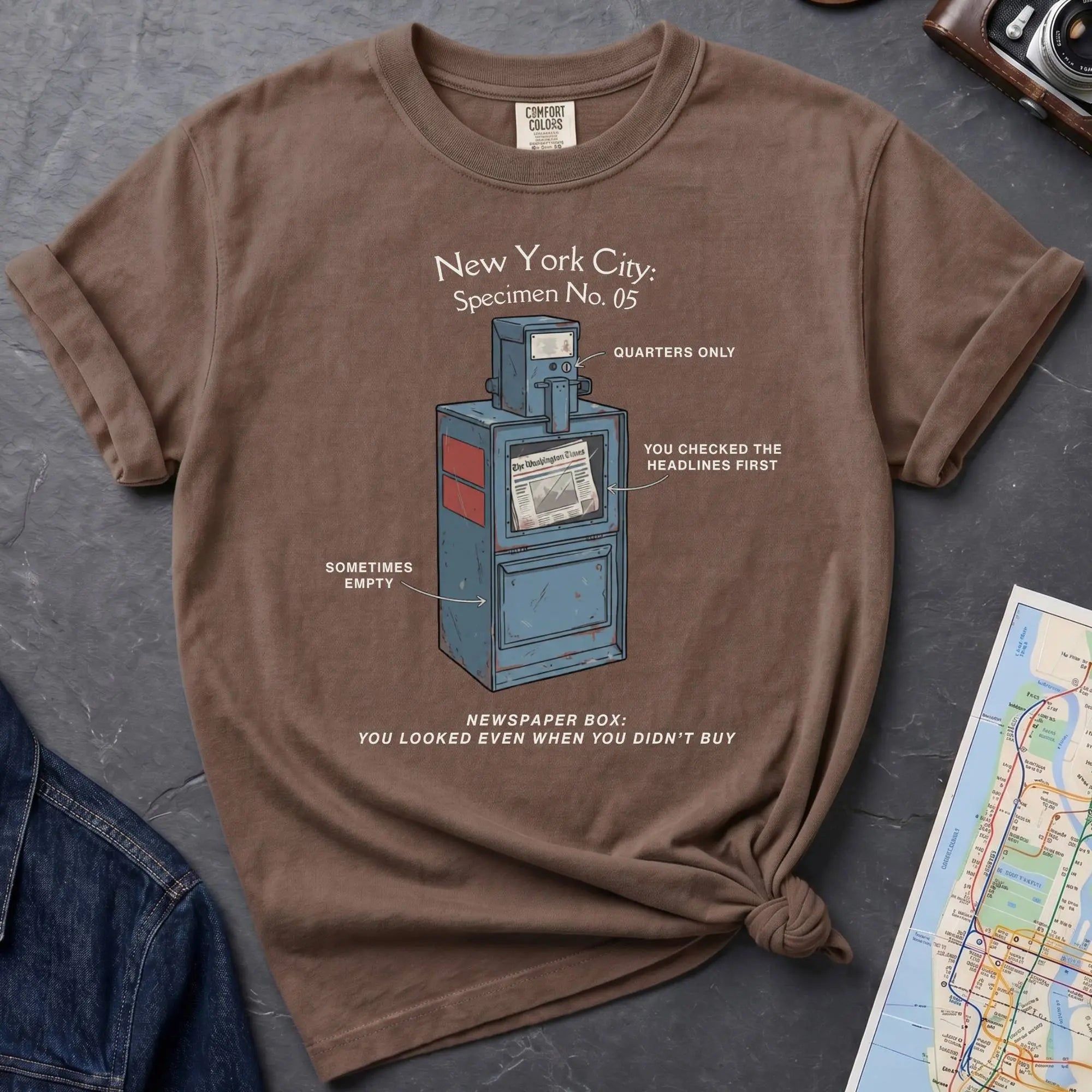 Brown t-shirt with 'Queens Est. 1683' graphic design on a gray surface with a map and jacket.