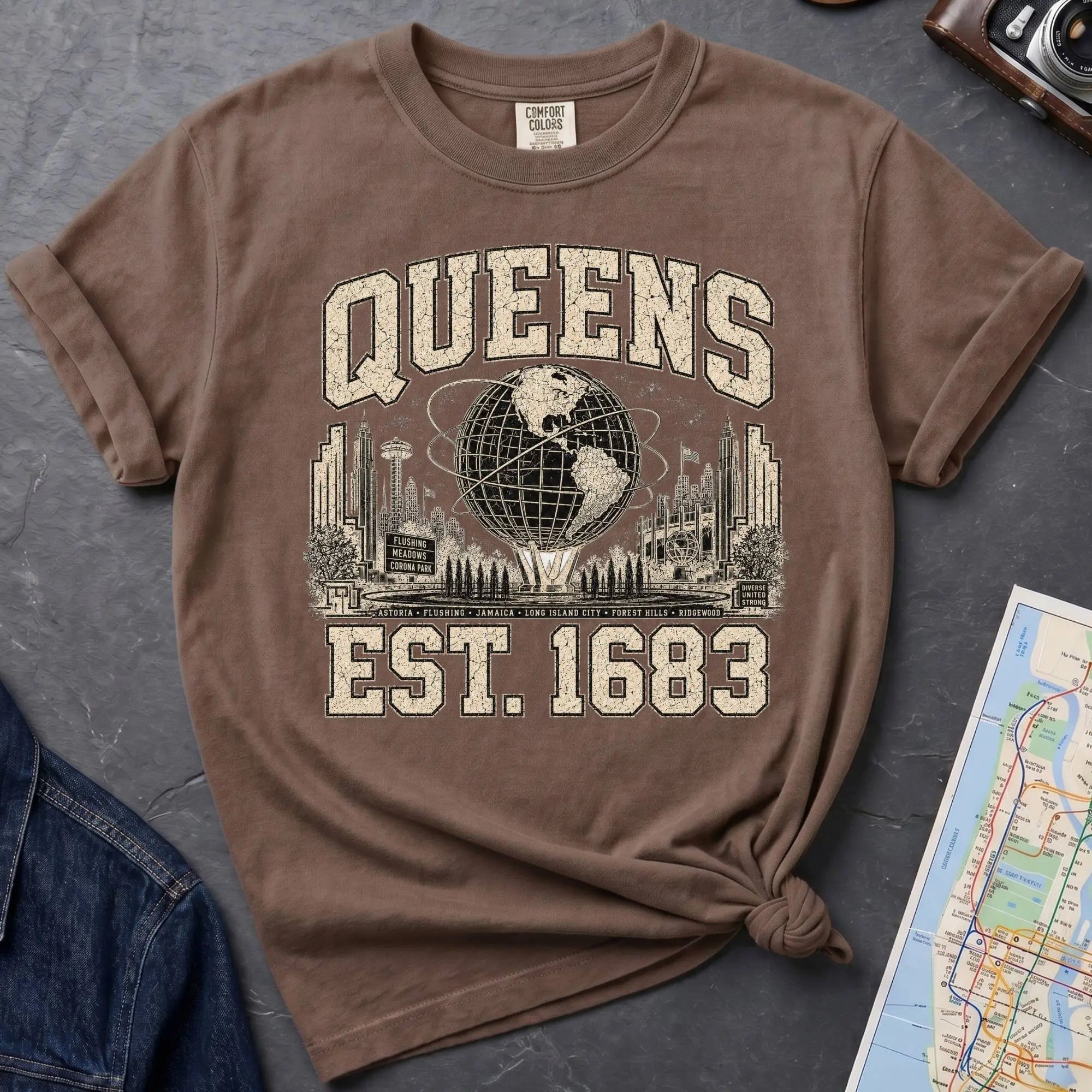 Brown t-shirt with 'Queens Est. 1683' graphic design on a dark surface with a denim jacket and a map.