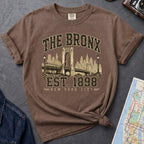 Brown t-shirt with 'The Bronx Est. 1898' design on a dark surface with a map and a denim jacket.