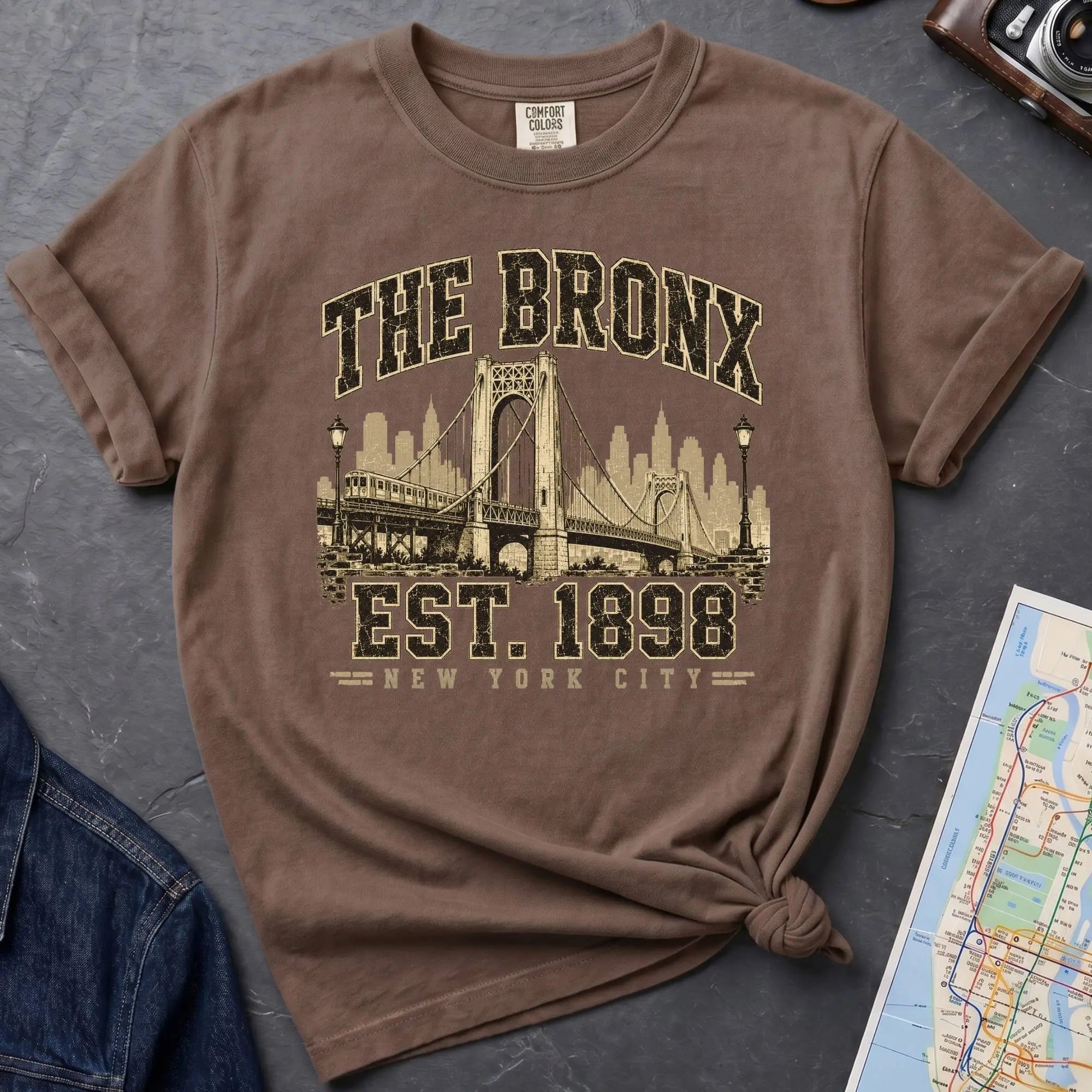 Brown t-shirt with 'The Bronx Est. 1898' design on a dark surface with a map and a denim jacket.