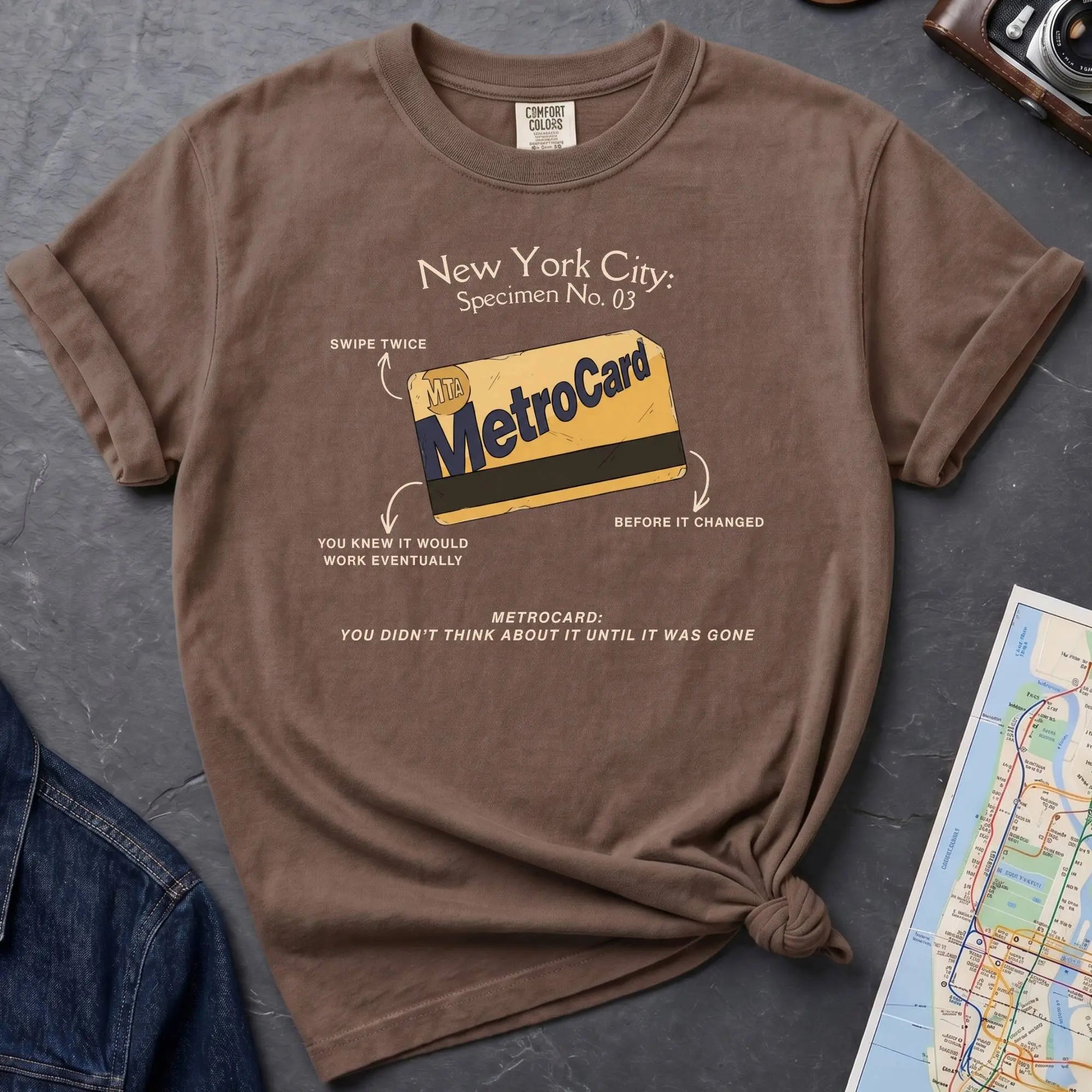 Brown t-shirt with New York City MetroCard graphic on a gray surface with a map and jacket.