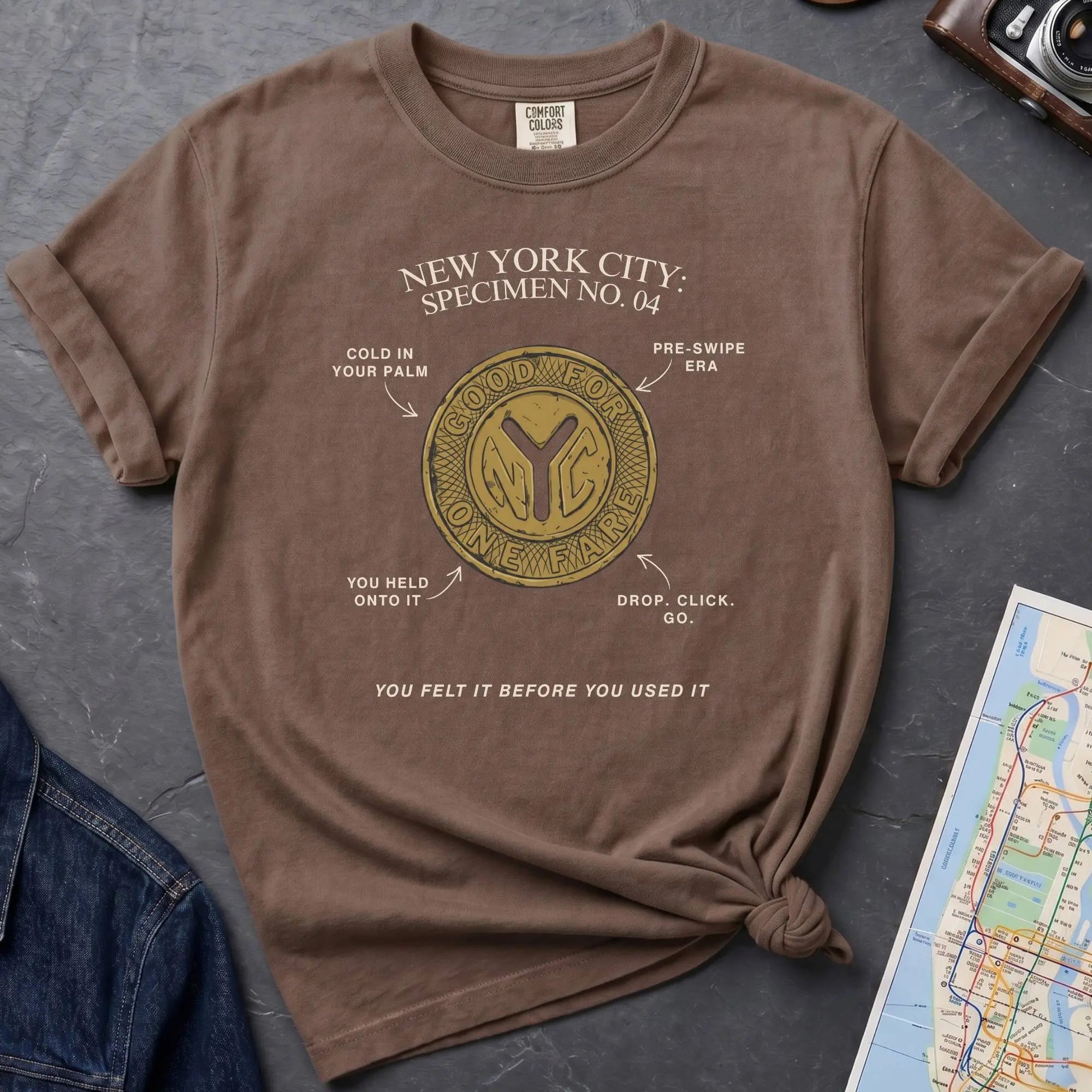 Brown t-shirt with New York City token design on a gray surface with a map and camera.