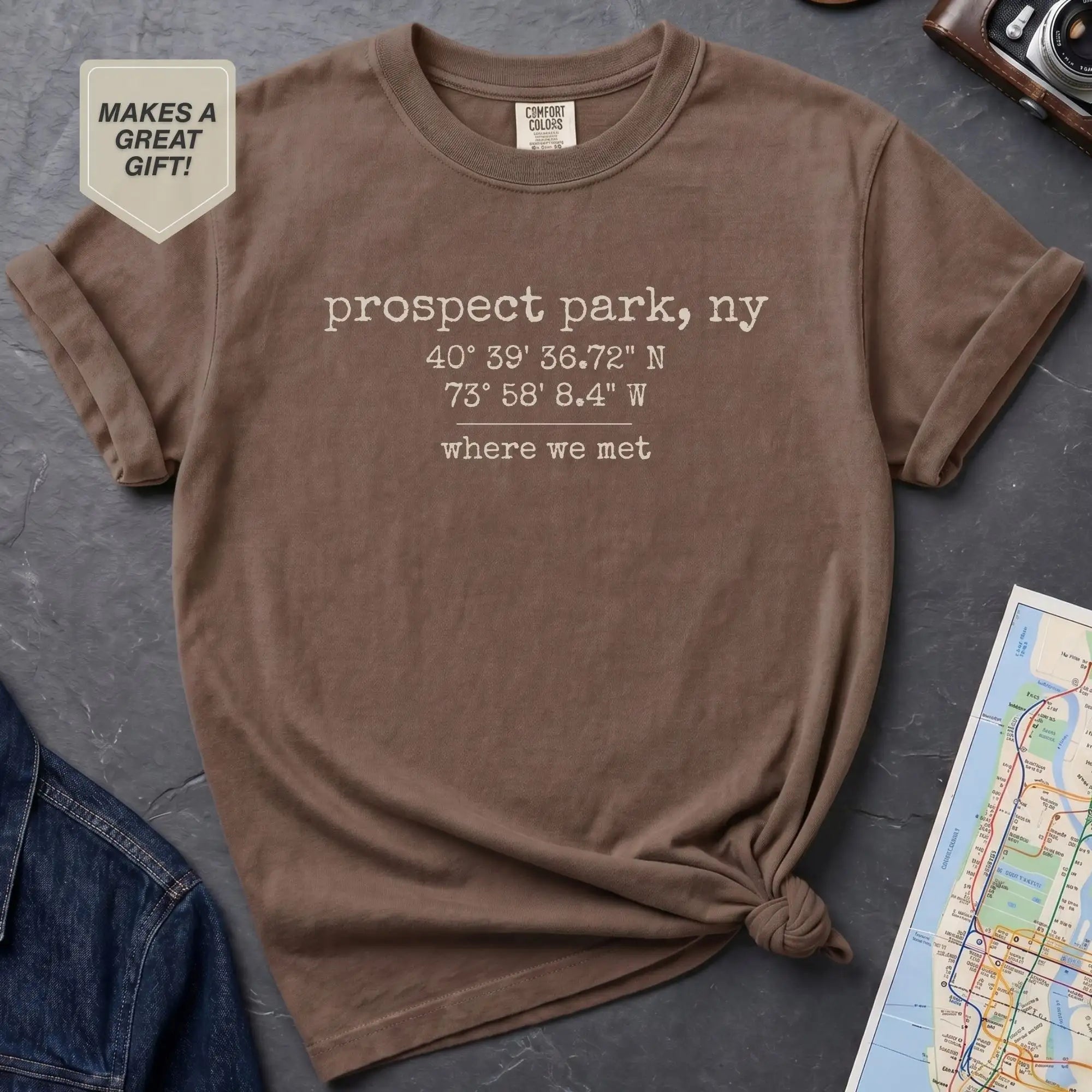 Brown t-shirt with text 'prospect park, ny' and coordinates on a gray surface with a map and jeans.