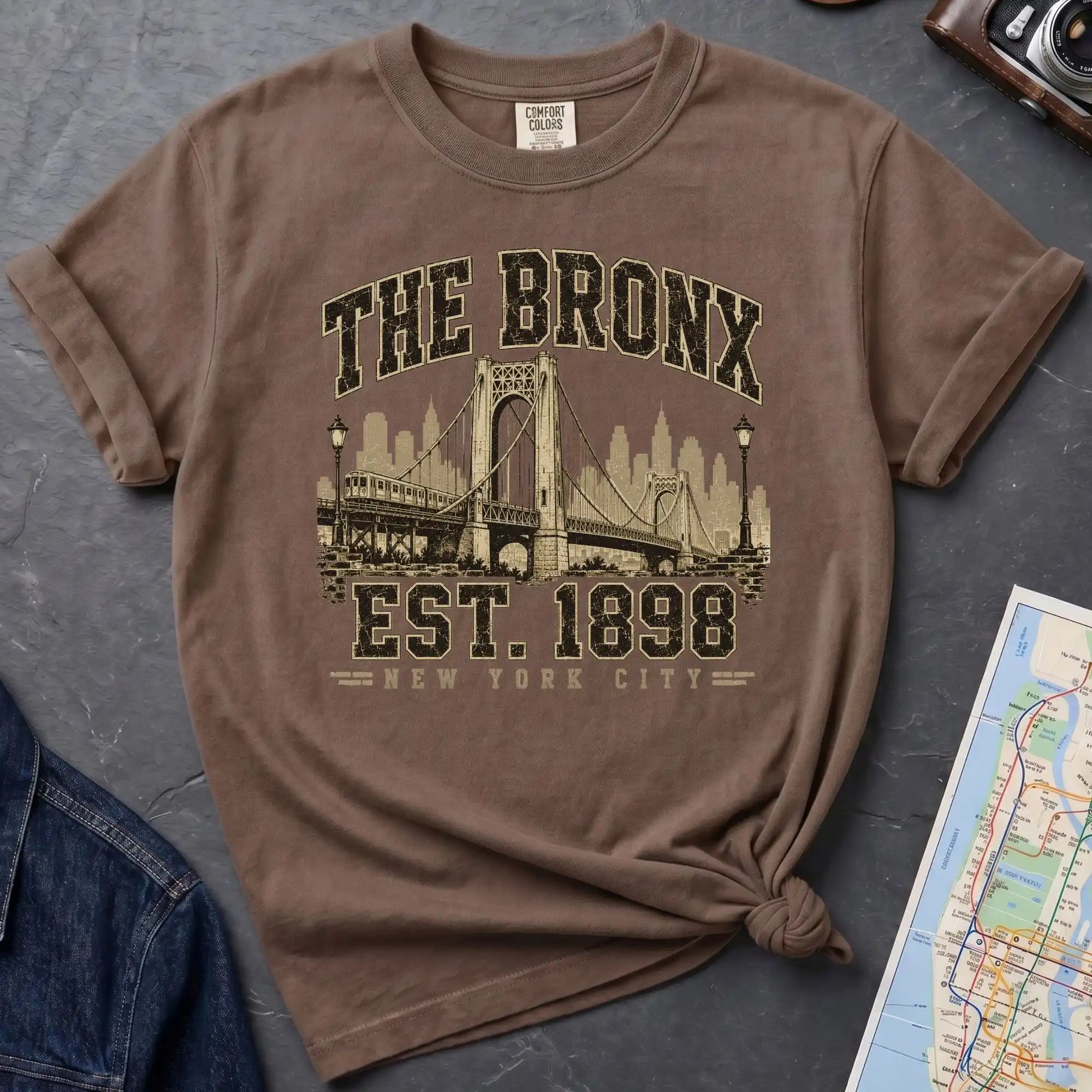 Brown t-shirt with 'The Bronx Est. 1898' design on a gray surface with a map and jacket.