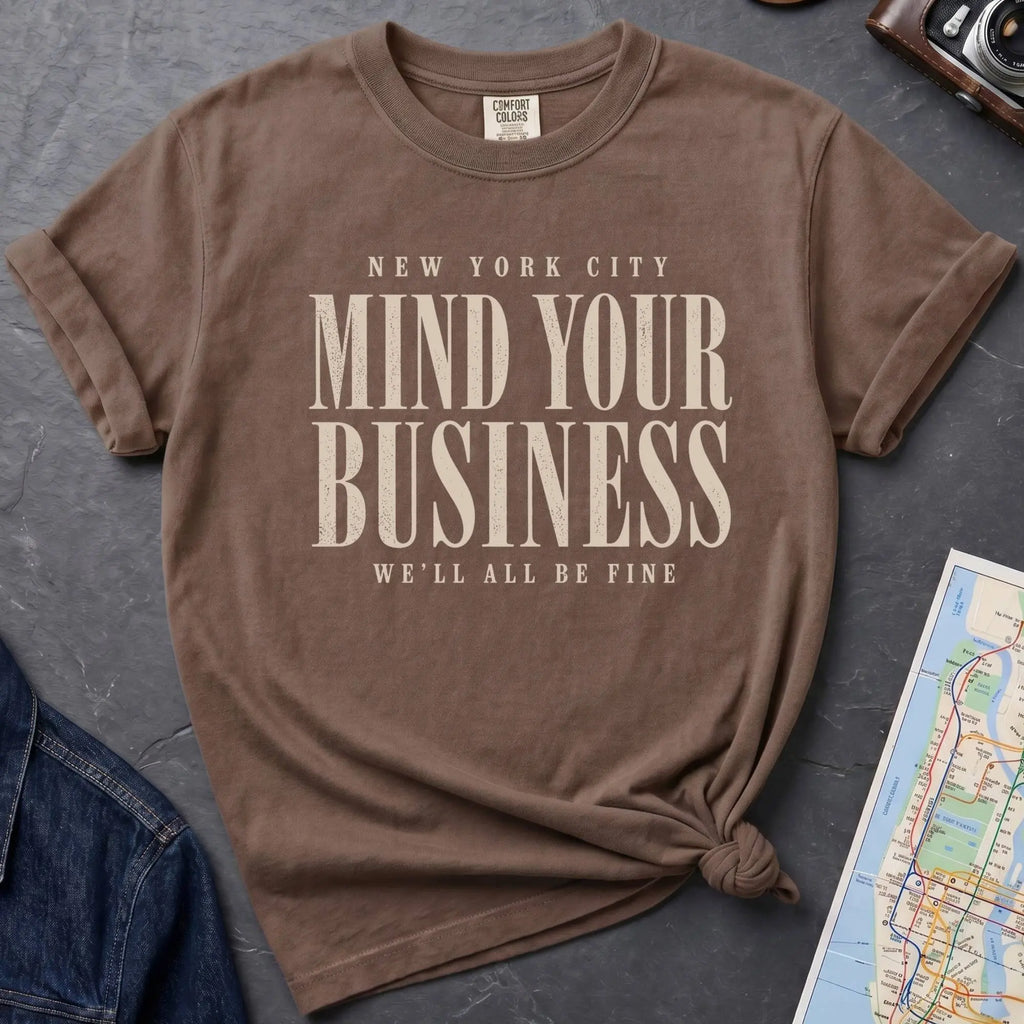 Brown t-shirt with 'New York City Mind Your Business We'll All Be Fine' text on a dark surface with a map and camera.