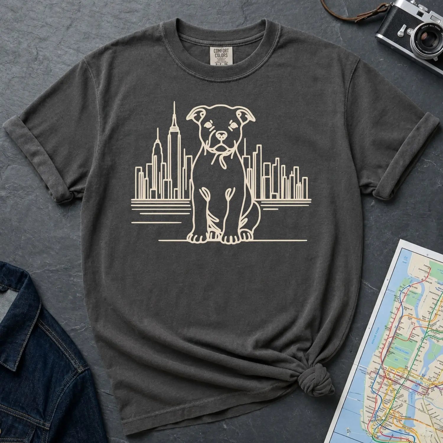 Pepper colored Comfort Colors t-shirt with a Pitbull dog and cityscape design on a gray surface with a denim jacket and map.