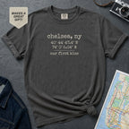 Pepper colored t-shirt with 'Chelsea, NY' coordinates and text on a gray surface with a map and camera.