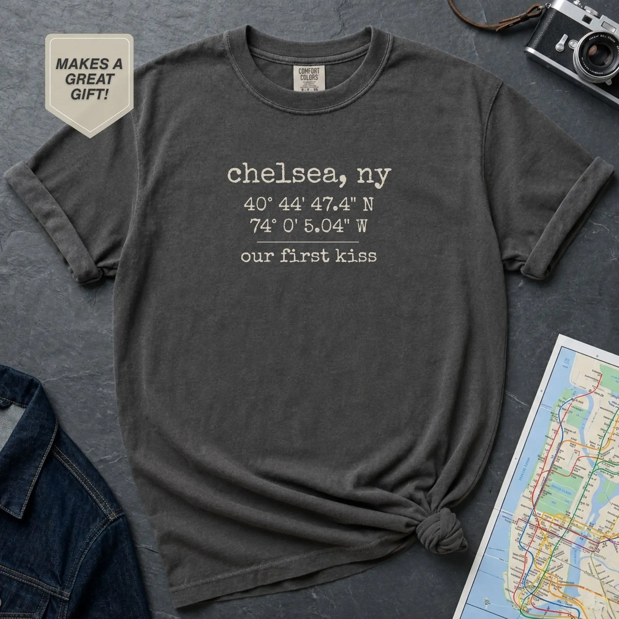 Pepper colored t-shirt with 'Chelsea, NY' coordinates and text on a gray surface with a map and camera.