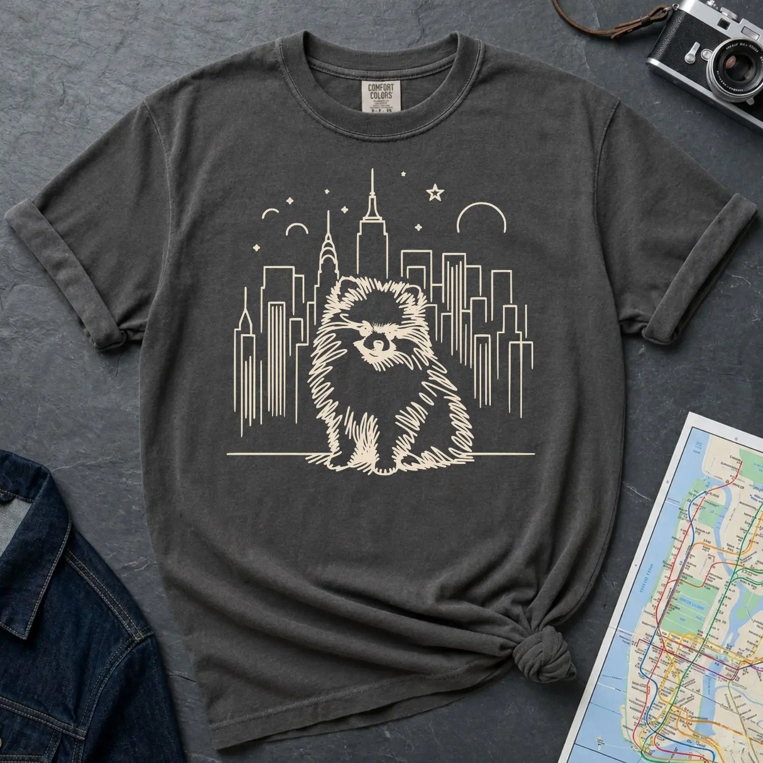 Pepper colored Comfort Colors t-shirt featuring a fluffy Pomeranian dog illustration in front of the New York City skyline on a dark surface with denim jacket and map.