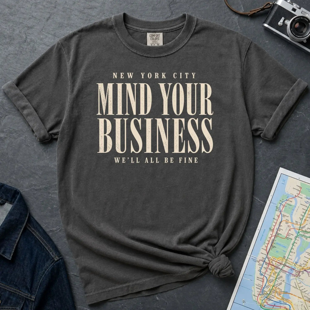 Pepper colored Comfort Colors t-shirt featuring bold New York City “Mind Your Business, We’ll All Be Fine” text in a vintage black typography design on a dark surface with a denim jacket and a map.