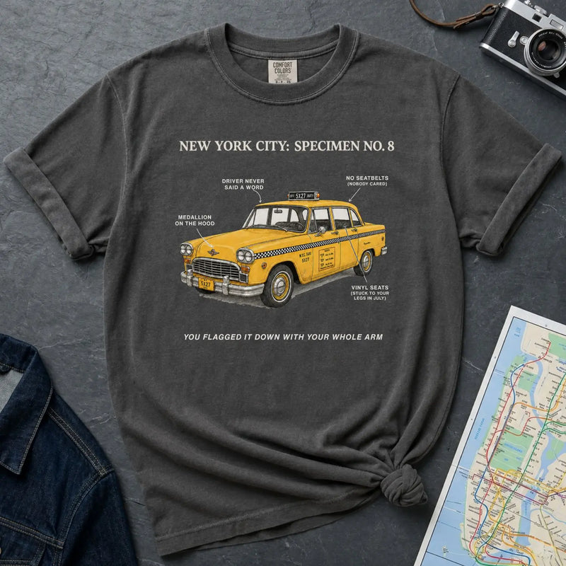 Pepper colored Comfort Colors t-shirt with a yellow taxi graphic and text on a dark surface with a map and camera.