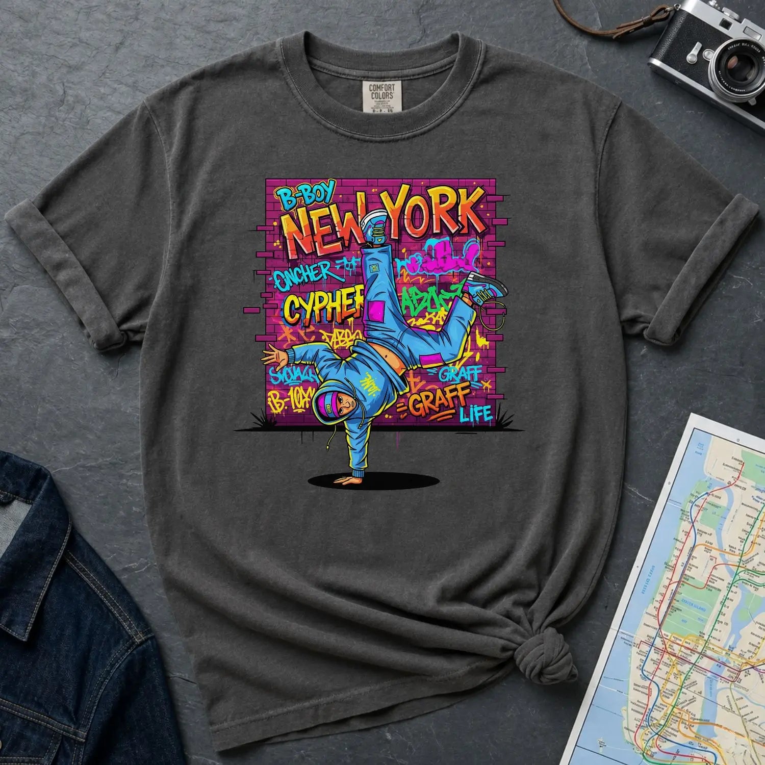 Pepper colored Comfort Colors t-shirt with a colorful graphic design featuring a breakdancer and text on a dark surface with a denim jacket and a map.