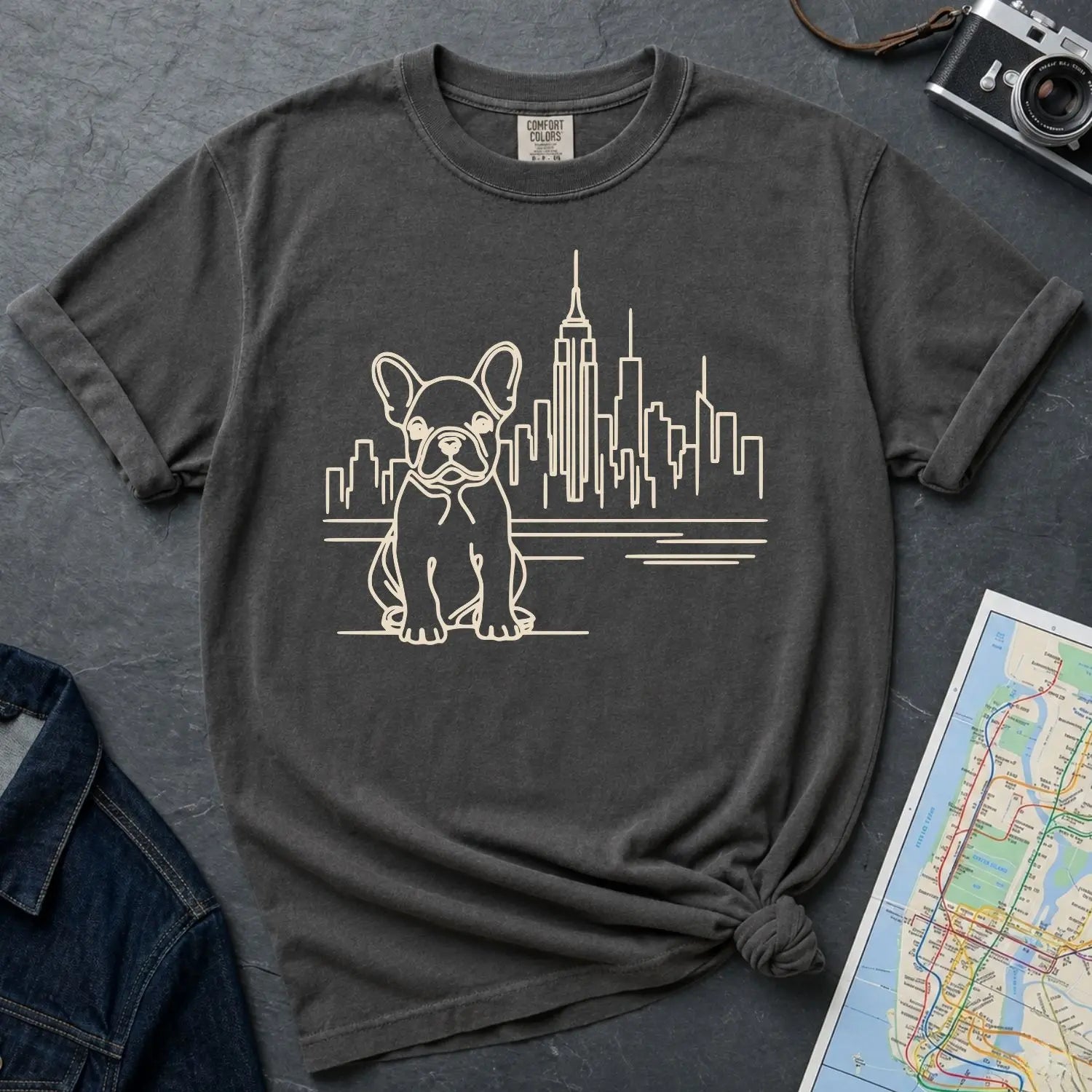 Pepper colored Comfort Colors t-shirt with a French Bulldog and cityscape design on a gray surface with a map and denim jacket.