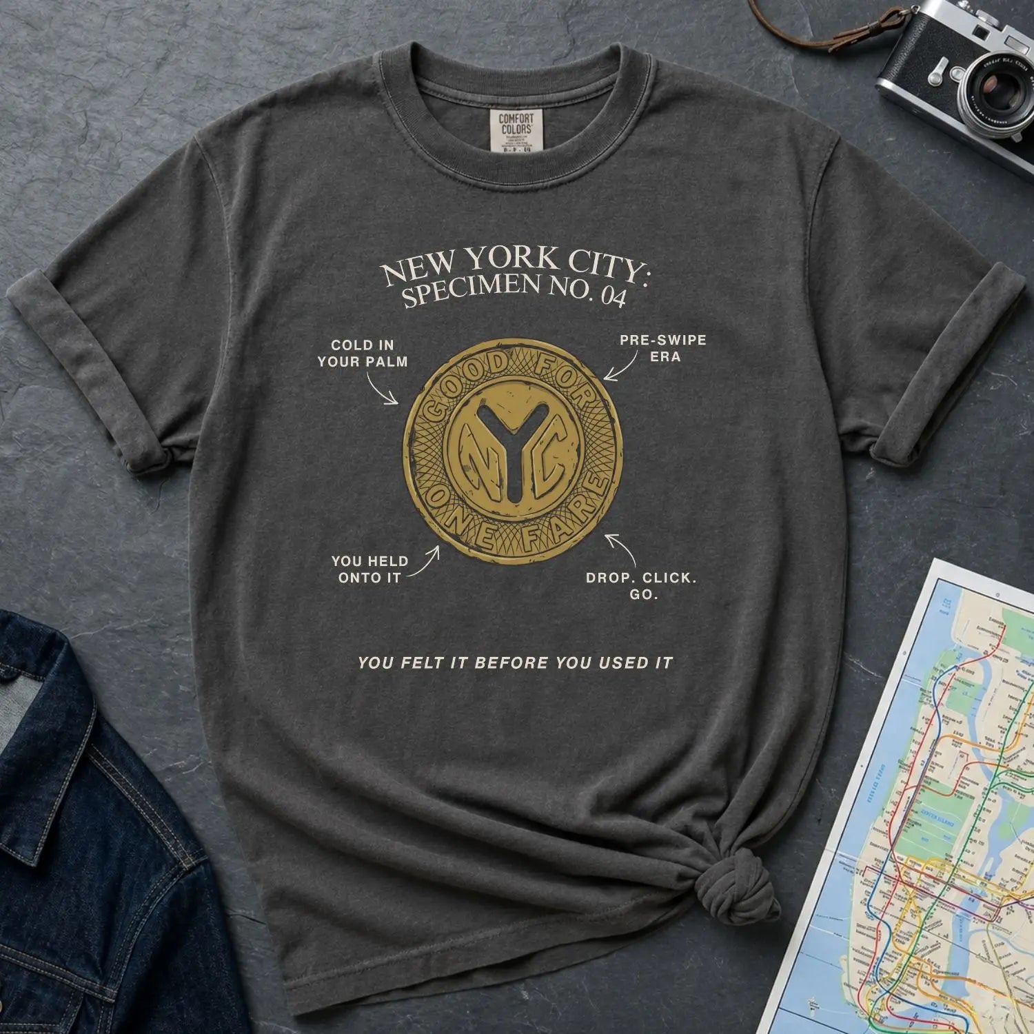 Pepper colored Comfort Colors t-shirt with New York City vintage token design on a gray surface with denim jacket and map.