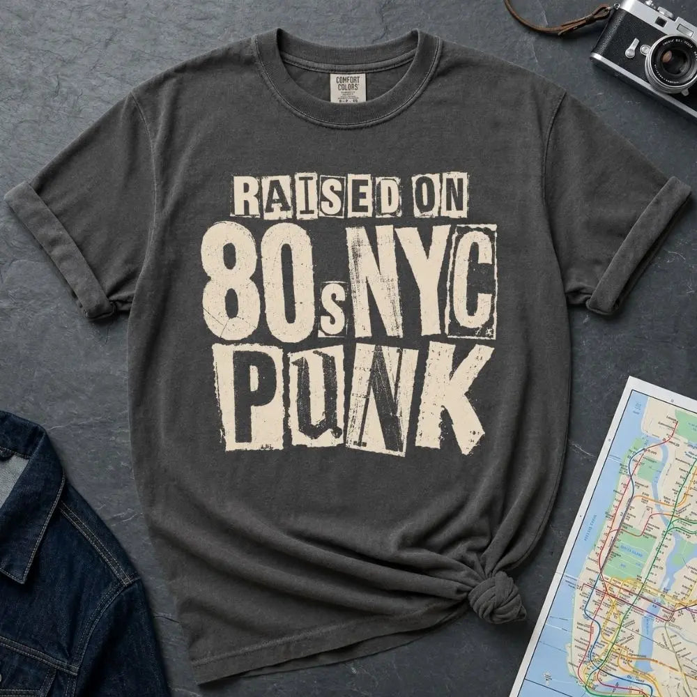 Pepper-colored Comfort Colors t-shirt with bold distressed ransom note style lettering reading "Raised on 80s NYC Punk" in mixed fonts and sizes, black ink on ivory, xerox zine aesthetic.
