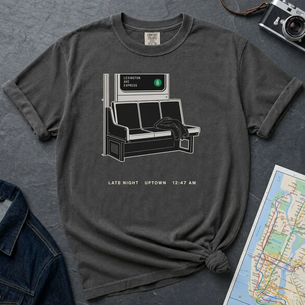 Pepper colored Comfort Colors t-shirt with a graphic of a subway bench and text, placed on a dark surface with a map and camera.