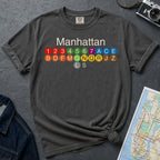 Pepper colored Comfort Colors t-shirt with Manhattan text and subway line design on a gray surface with a map and camera.
