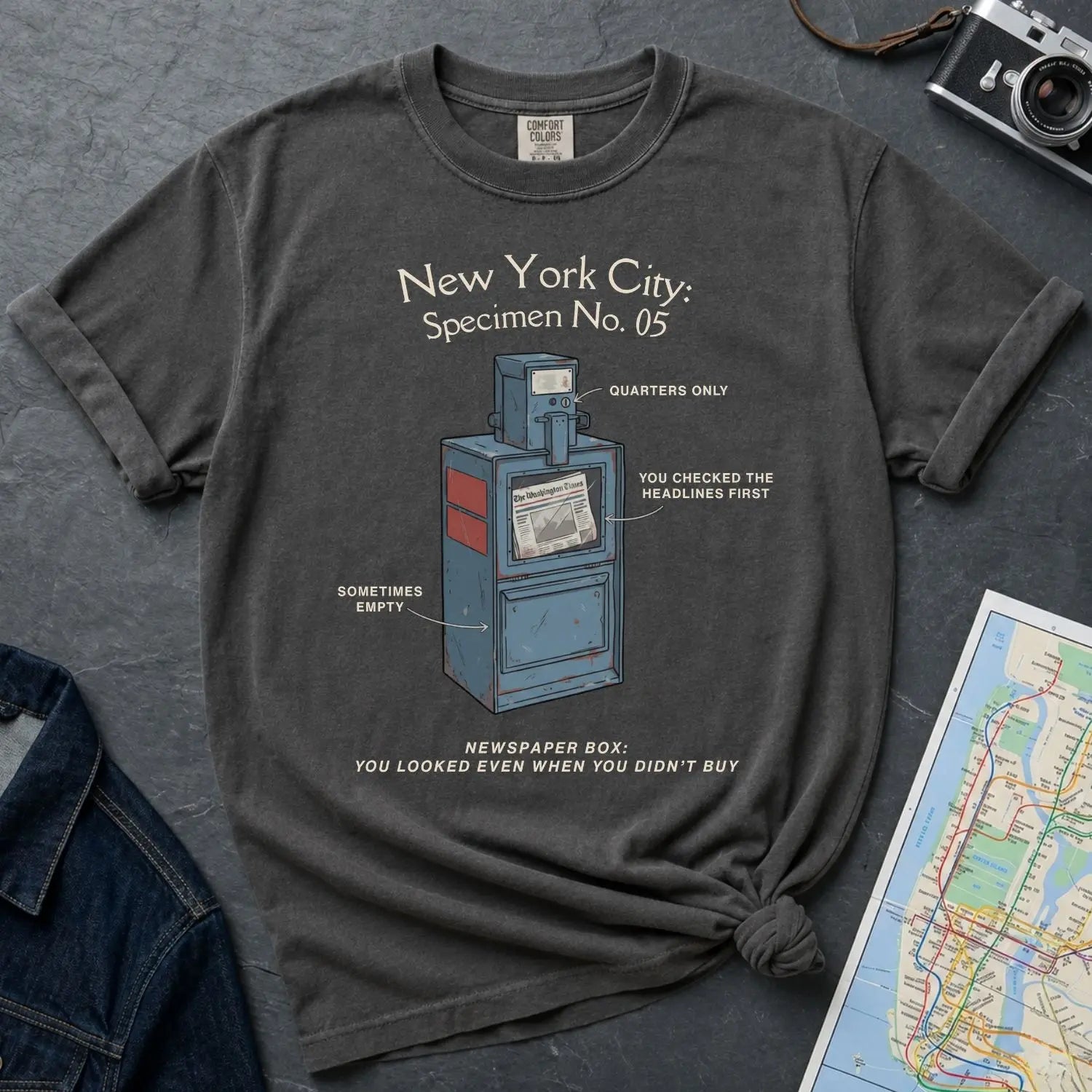 Pepper colored Comfort Colors t-shirt with a newspaper box graphic and text on a textured surface with a camera and map.
