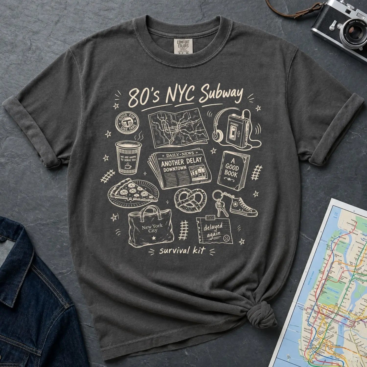 Pepper colored Comfort Colors t-shirt with '80s NYC Subway Survival Kit' graphic design on a textured surface with a map and camera.