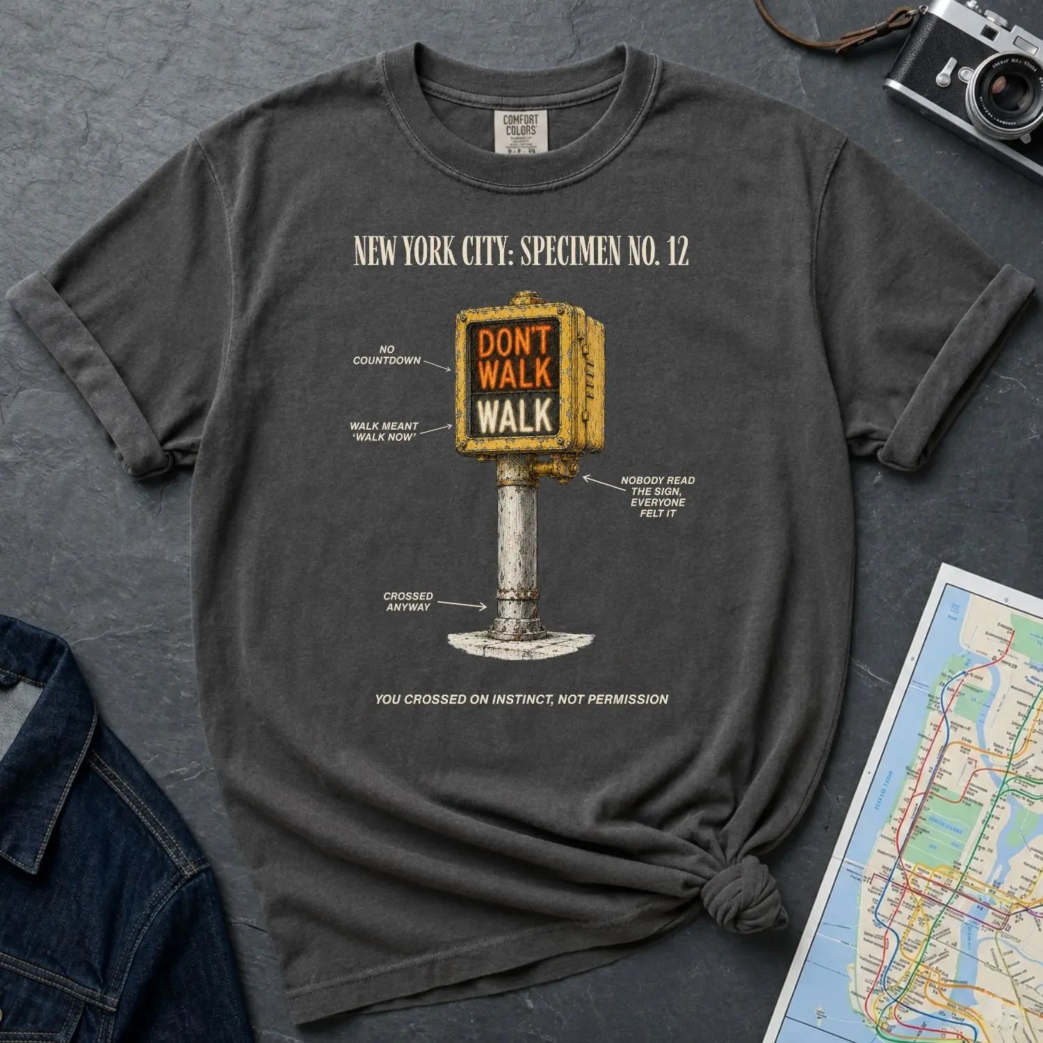 Pepper colored Comfort Colors t-shirt with a graphic design of a vintage intersection crossing sign and text, placed on a dark surface with a denim jacket and a map.