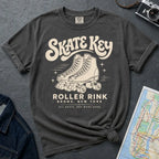 A pepper-colored Comfort Colors t-shirt featuring a cream-colored retro design with roller skates and the text 'Skate Key Roller Rink Bronx, New York'. The t-shirt is styled in a flat-lay composition on a textured gray background, surrounded by props including a denim jacket, a vintage camera, and a map.