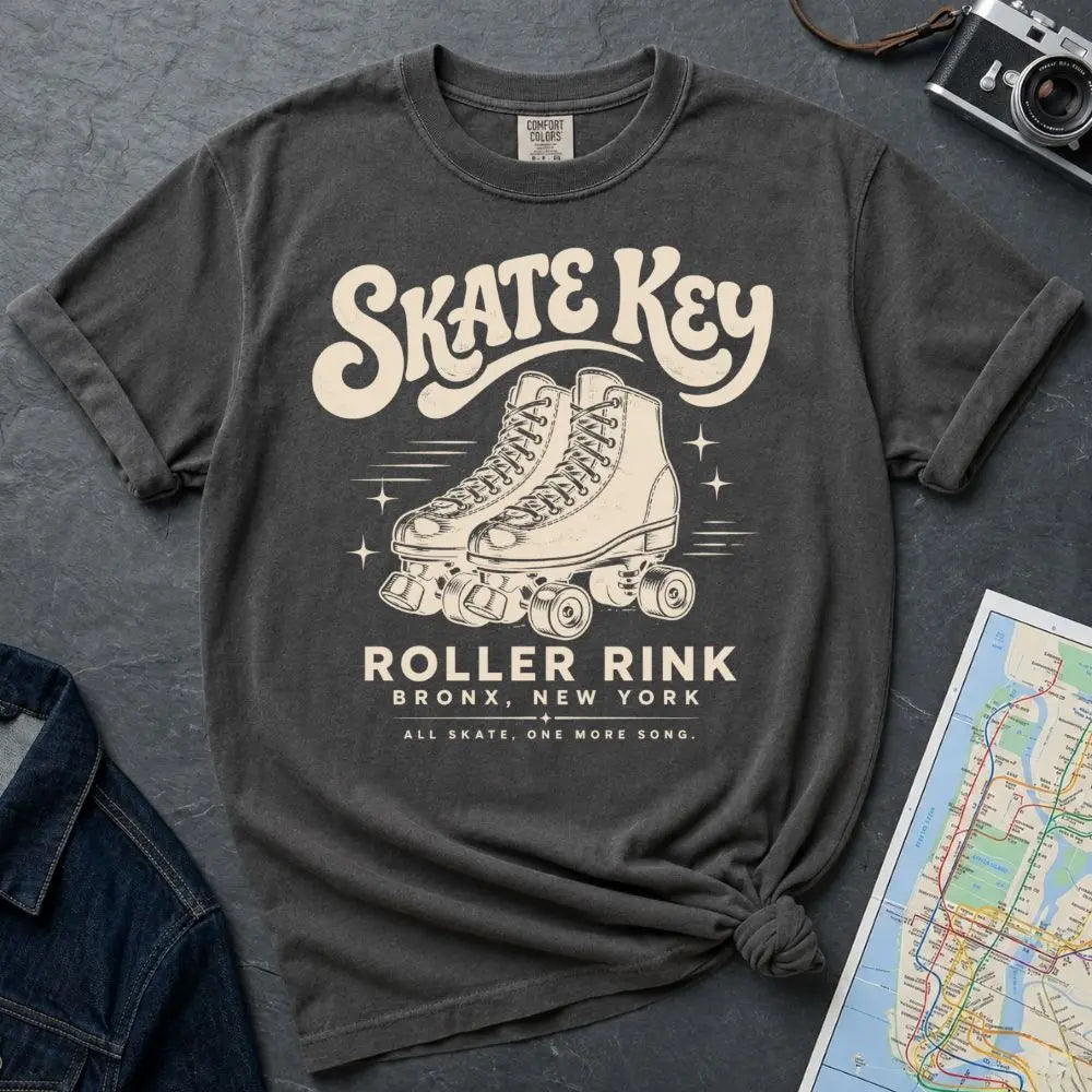 A pepper-colored Comfort Colors t-shirt featuring a cream-colored retro design with roller skates and the text 'Skate Key Roller Rink Bronx, New York'. The t-shirt is styled in a flat-lay composition on a textured gray background, surrounded by props including a denim jacket, a vintage camera, and a map.